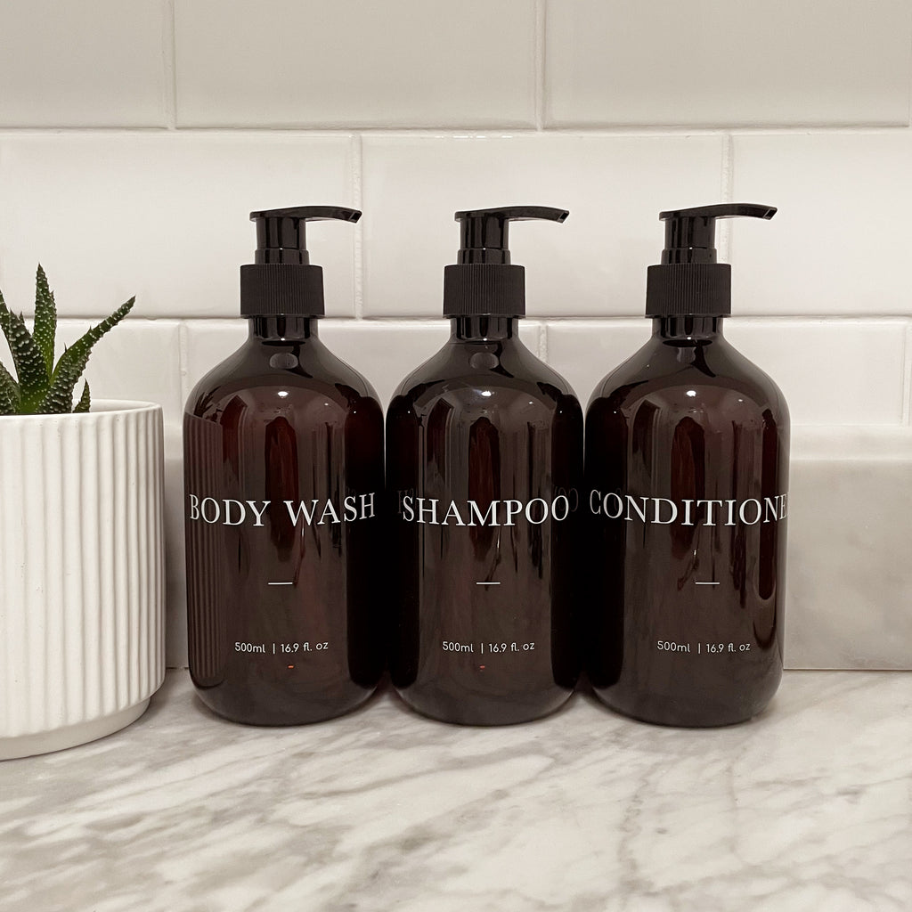 Three brown bathroom pump bottles labeled 'Body Wash', 'Shampoo', and 'Conditioner'. Elegant refillable bathroom bottles with customisable text printed on,
