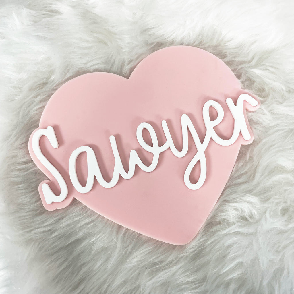 Pink heart-shaped baby name sign with the name 'Sawyer' on a fluffy white blanket. Cute newborn baby photo props and nursery decor.